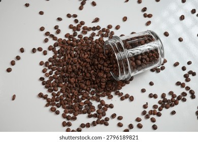 Top view composition of roasted coffee beans spilled from a glass jar on white background.