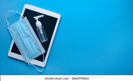 Top view of computer tablet, face mask and a bottle of hand sanitizer isolated on a blue background with copy space.