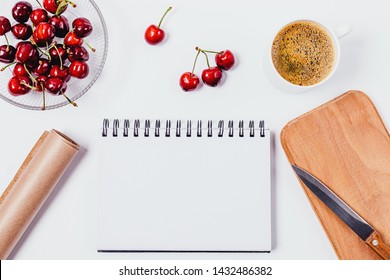 Top View Cooking Template For Text On An Empty Notepad Sheet Next To Wooden Board And Knife, Baking Paper, Bowl Of Fresh Cherries And Cup Of Coffee On White Kitchen Table.