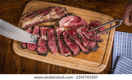 Top view of cote de boeuf steak being prepared on wooden board with knife and meat serving fork on wooden table in restaurant