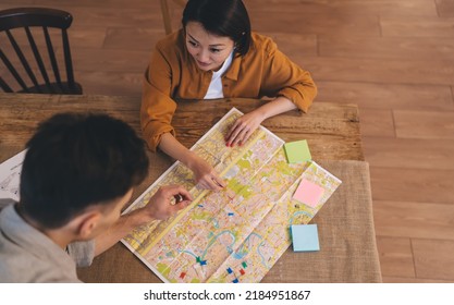 Top View Of Couple Choosing Famous Places On City Map At Table At Home. Asian Woman Looking On Boyfriend. Persons Planning Vacation Or Journey. Wooden Desk With Adhesive Paper And Notice Board Pins