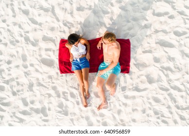 Top View Of Couple Lying On White Sand Beach Taking A Sunbath In Summer