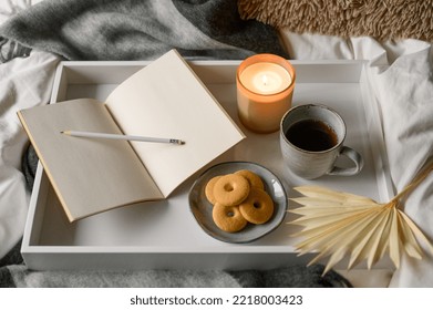 Top View Of Cozy Autumn Composition Burning Aroma Candle, Open Notebook, Pencil, Cup Of Coffee  On White Wooden Tray With Gray Blanket In Background On Comfy Bed