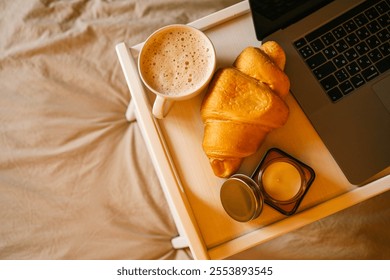 Top view of a cozy breakfast setup with a frothy cup of coffee, a golden croissant, and a small jar of honey placed on a white tray beside a laptop on a soft beige bed. - Powered by Shutterstock