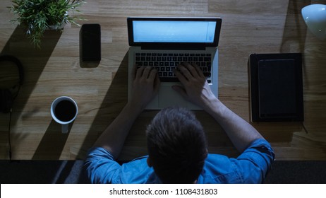 Top View Of Creative Designer Working On His Laptop At Night. His Table Is Illuminated By Cold Blue Light From Outside.