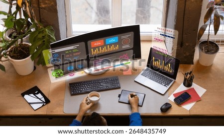 Top view of creative digital analyst working at a desk with curved monitor, laptop, and holographic infographics. Concept of smart work environments, creative economy workspace