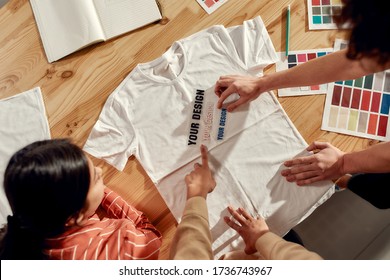 Top view of creative people trying on stickers with text, while discussing logo and design of T-shirt. Young man and women working together at custom T-shirt, clothing printing company. Cropped shot
