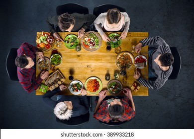Top view creative photo of friends sitting at wooden vintage table. Friends of six having dinner. They with plates full of meal and glasses with drinks. There is free space in the middle of table