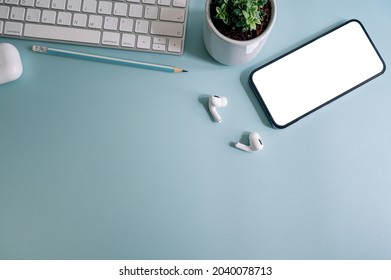 Top view creative workspace with computer keyboard, blank screen smartphone, earphone and plant on light blue background.