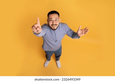 Top view of creative young asian guy in casual outfit pointing finger up while standing on yellow studio background, cheerful male having nice idea, showing eureka gesture, full length, copy space