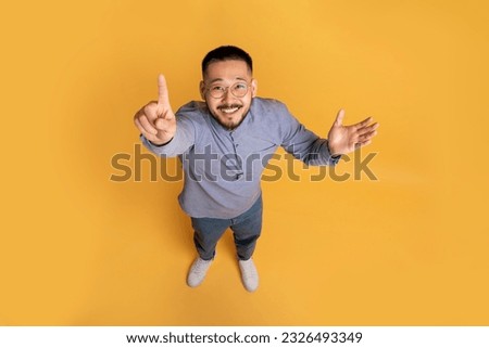 Top view of creative young asian guy in casual outfit pointing finger up while standing on yellow studio background, cheerful male having nice idea, showing eureka gesture, full length, copy space