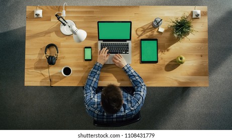 Top View Of A Creative Young Man Sitting At His Desk Typing On Laptop Computer Beside Lies Smartphone And Tablet Computer, All Electronic Devices Have Green Mock-up Screen.