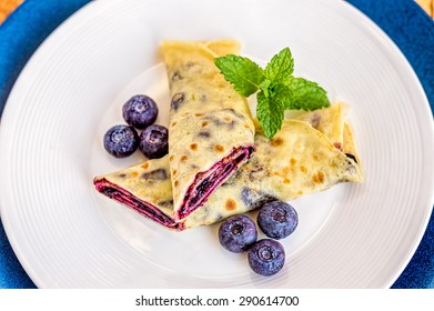 Top View Of Crepe With Blueberry Jam Filling Cut In Half And Decorated With Mint Leaves And Frozen Blueberries.