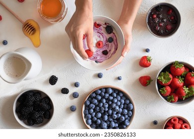 Top view of crop woman hands preparing breakfast with bowl with natural yogurt and raspberries, blueberries, strawberries and blackberries near bowls with honey and different fresh fruits on table स्टॉक फोटो