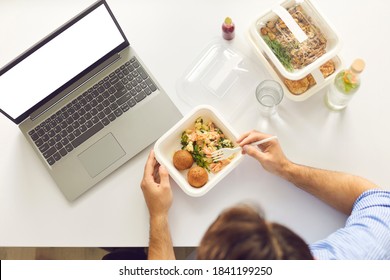 Top view of cropped busy worker sitting at office desk with laptop computer with blank white mockup copyspace screen, enjoying lunch break and eating healthy meal ordered on food delivery website