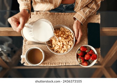 Top view cropped photo of a woman preparing herself breakfast while pouring milk into a plate of cereal, strawberries and a cup of coffee standing nearby. Breakfast woman in cozy kitchen. - Powered by Shutterstock