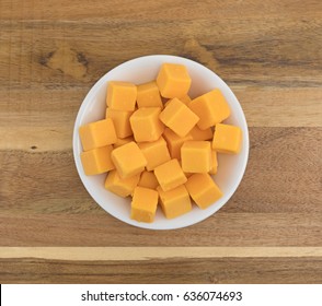 Top View Of Cubed Mild Cheddar Cheese In A White Bowl Atop An Old Wood Counter Top.