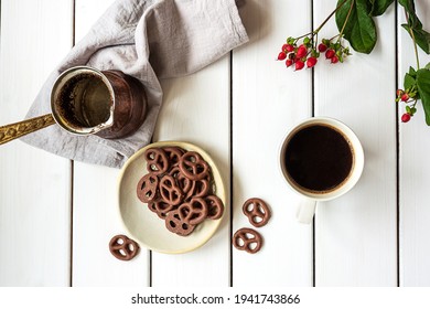 Top view of a cup of coffee, coffee pot and chocolate-covered pretzels on a white wooden background. Breakfast or coffee break composition.