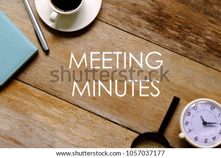 Top view of a cup of coffee,notebook,pen,magnifying glass and clock on wooden background written with 'MEETING MINUTES'.