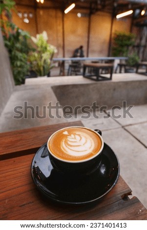 top view of a cup of hot cappuccino coffee with heart-shape latte art on black saucer on stool with blurred coffee shop interior. morning breakfast