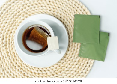 A top view of a cup of tea placed on a round woven coaster. Product labels are on the rim of the cup without a logo. Two unpatterned green paper packets for herbal tea bags on woven placemats. - Powered by Shutterstock