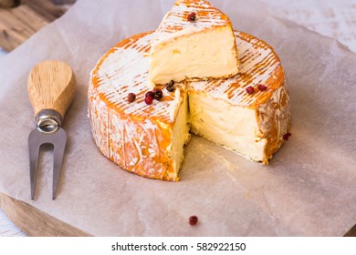 Top view of cut slice of French or German soft cheese with orange rind with mold, creamy texture, red pepper corns, fork,on parchment paper, wood cutting board  - Powered by Shutterstock