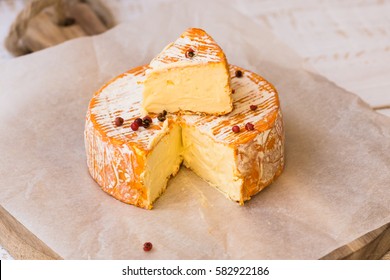 Top view of cut slice of French or German soft cheese with orange rind with mold, creamy texture, red pepper corns, waxed paper, wood cutting board - Powered by Shutterstock