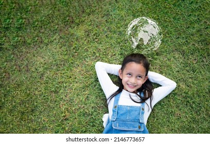 Top View Of Cute Asian Little Girl On The Green Grass And She Is Happy Thinking About Love, Friend, Freedom, Peaceful, Diversity And Connectivity Of People Around The World And Save The World Concept.