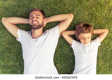 Top view of cute little boy and his handsome young dad smiling while lying with hands behind head and closed eyes on the grass