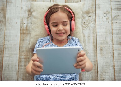 Top view of cute little girl in headphones listening to music using a tablet and smiling while lying on wooden floor - Powered by Shutterstock