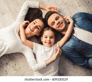 Top view of cute little girl and her beautiful young parents looking at camera and smiling while lying on the floor at home