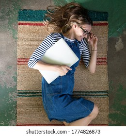 La vue de dessus d'une femme mignonne dans des lunettes porte des salopettes de jeans bleus, allongée sur un tapis rouge sur le sol en béton, tenant son ordinateur portable et se reposant après le travail. 