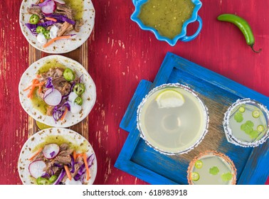 Top View Of Cutting Board With Pork Verde Tacos And Glasses Of Margaritas On A Blue Tray.