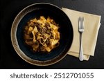 Top view of Dark bowl with pappardelle with wild boar ragout sauce with napkin and fork on black stone