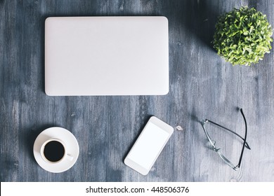 Top View Of Dark Wooden Office Desktop With Closed Laptop, Blank White Smartphone, Plant, Glasses And Coffee Cup. Mock Up