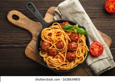 Top view of delicious pasta with meatballs, tomato sauce and fresh basil in cast iron rustic vintage pan served on cutting board, wooden background. Tasty homemade meatballs spaghetti 