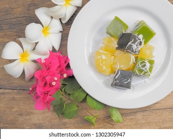 Top View Of Delicious Thai Dessert Khanom Piek Poon (Burnt Coconut Jelly), Khanom Chun (Thai Jello) And Khanom Man (Coconut Cassava Cake) In White Plate With Flowers On Wood Texture Background.