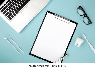 Top View Of Dentist Table With Blank Paper On Clipboard, Pen, Computer Laptop, Eyeglasses, Tooth Model, Toothbrush, Dental Explorer Probe And Dentist Mirror. 