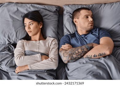 top view of depressed interracial couple lying down with folded arms and looking away on bed. Translation of tattoo: be faithful to the one who is faithful to you - Powered by Shutterstock