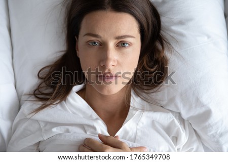 Top view of depressed upset young woman lie relax on white pillow linen sheets feel distressed lonely, sad millennial female rest in home bedroom suffer from depression or insomnia, having problems