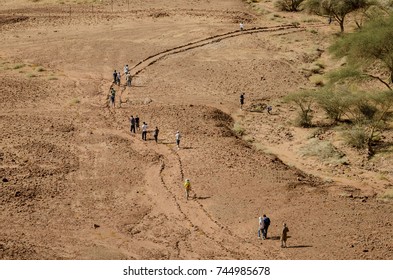 Top View Of A Desert Landscape While People Walk Down The Road. Some Green Acacia Trees Frame The Composition On The Right Side.