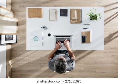 Top View, Designer Sitting At Desk And Working On His Laptop, His Table Is Perfectly Tidy, The Sun Casts Graphics Shadows On The Wood Floor