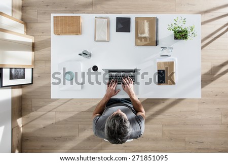 top view, designer sitting at desk and working on his laptop, his table is perfectly tidy, the sun casts graphics shadows on the wood floor