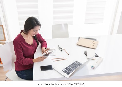 Top View Of An Designer Working Home On New Ideas A Dark Hair Braided Woman Is Sitting At A White Table In Casual Clothes, She Is Looking At Her Tablet And With Her Laptop And Some Pencils Next To Her