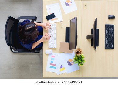 Top View Of The Desk In The Office Of An Asian Woman Who Is Sitting In Front Of The Computer And Sheet Of Statistics And Graphs Information. By Sitting Alone Which The Opposite Table Has No People Com