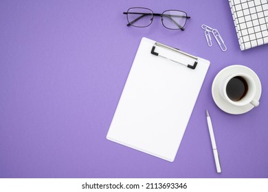 Top view desk with office supplies on purple background. Notepad, glasses, pen, coffee cup, paper clips on purple background. Flat lay
