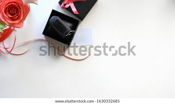 Top\
view Digital car key putting inside the black gift box with red\
ribbon,bouquet of roses and wish card on the white desk as\
background. Surprising Valentine\'s Day gift\
concept.