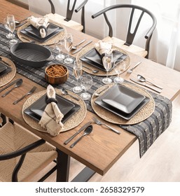 Top view of a dining table set for a meal, featuring modern black dinnerware on rustic woven placemats, complemented by elegant glassware, metallic utensils, and a stylish wooden table, well-lit room.