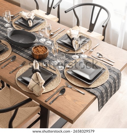 Top view of a dining table set for a meal, featuring modern black dinnerware on rustic woven placemats, complemented by elegant glassware, metallic utensils, and a stylish wooden table, well-lit room.