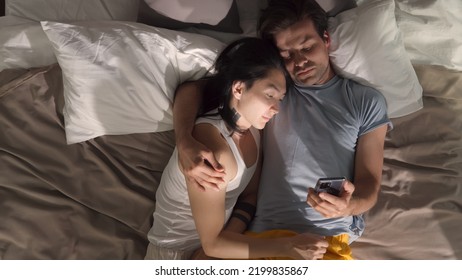 Top View Of Diverse Gay Couple Looking At The Phone And Talking On Bed. Asian And Caucasian Homosexual Partners Lie In Bed Hugging And Using Smartphone